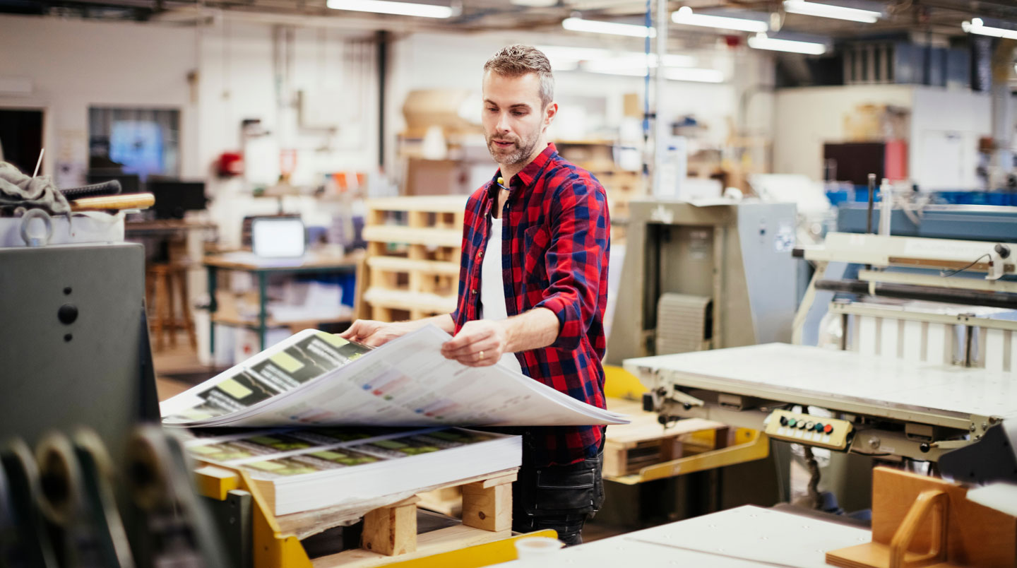 Conseillère d'une agence d'impression remettant un carton de commande à un client au comptoir d'accueil, dans un espace moderne lumineux avec signalétique en français et étagères d'échantillons en arrière-plan